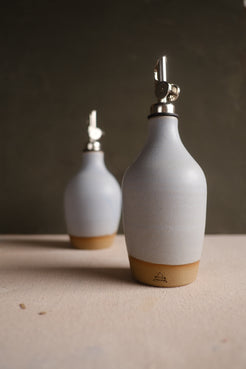 Wander Oil Bottle in white glaze with exposed clay base and stamp, beside a second bottle on canvas.