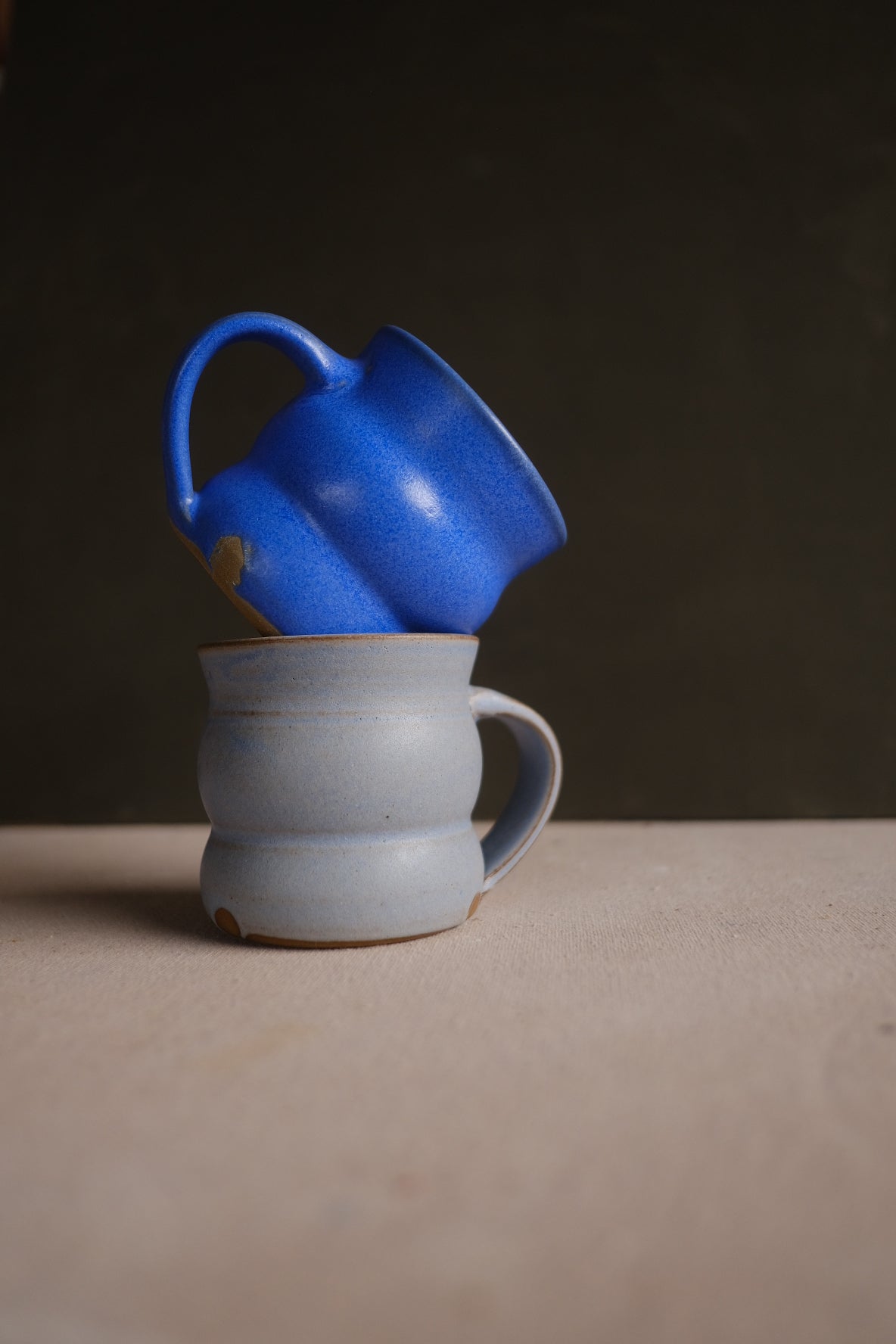 The Wave Mug in cobalt blue resting tilted atop an upright white glazed stoneware mug with unglazed foot.