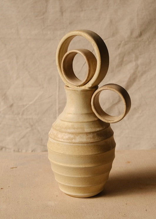Within Vase with concentric ring stopper, horizontal ridges, and round side handle pointing right on pale fabric.