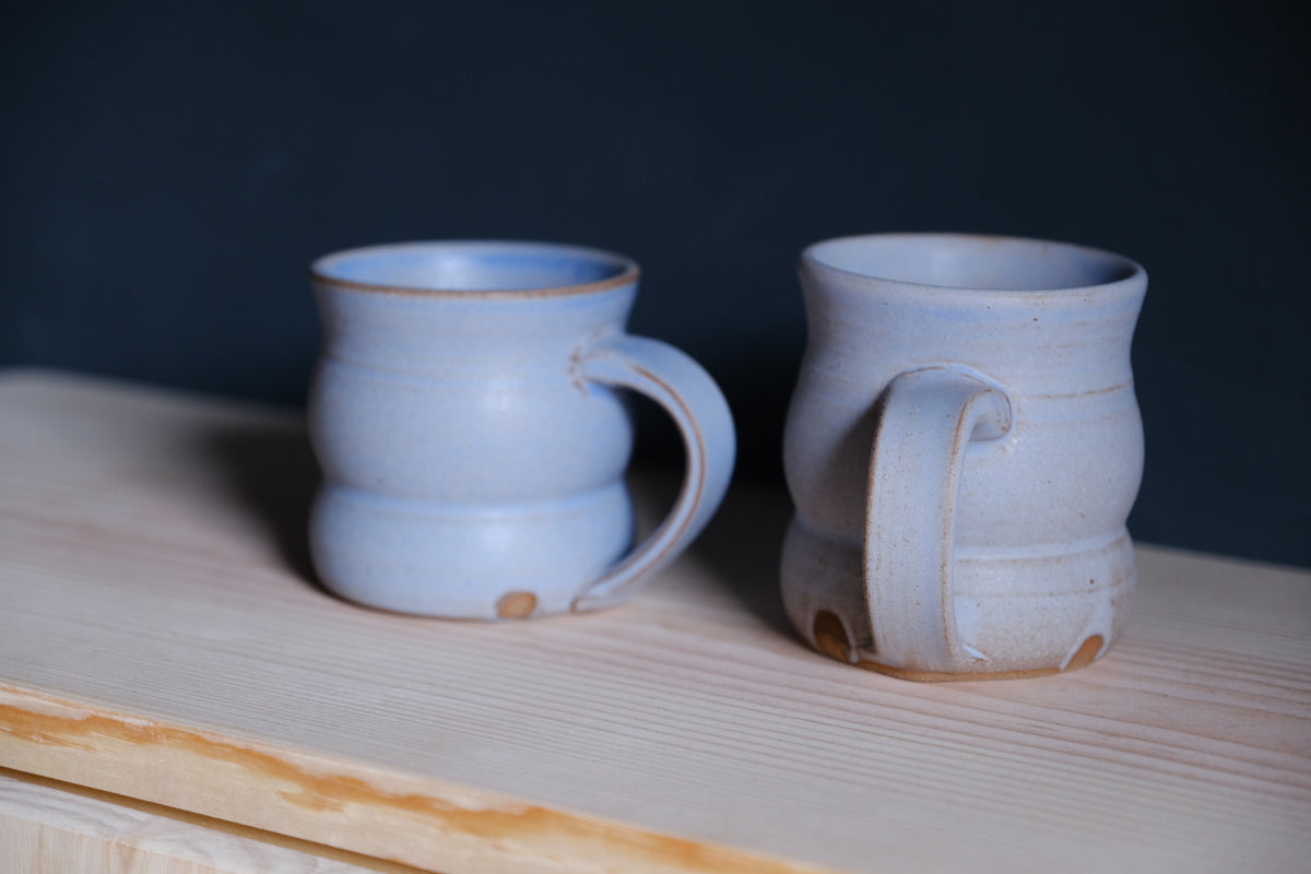 The Wave Mug pair in misty periwinkle glaze on an untreated wood shelf showing wide pulled handles.