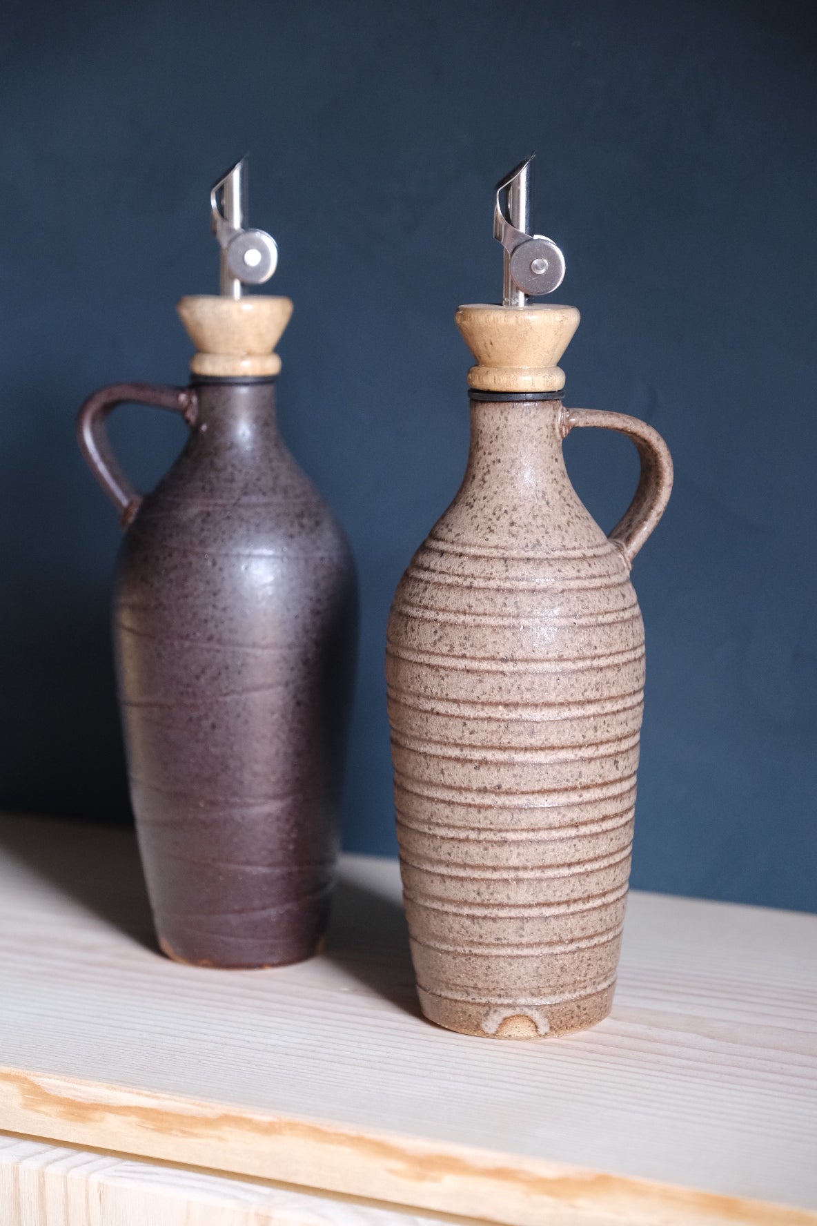 Tejas Oil Bottle set of two on a wooden shelf, dark brown and light speckled stoneware, metal spouts with wood collars.