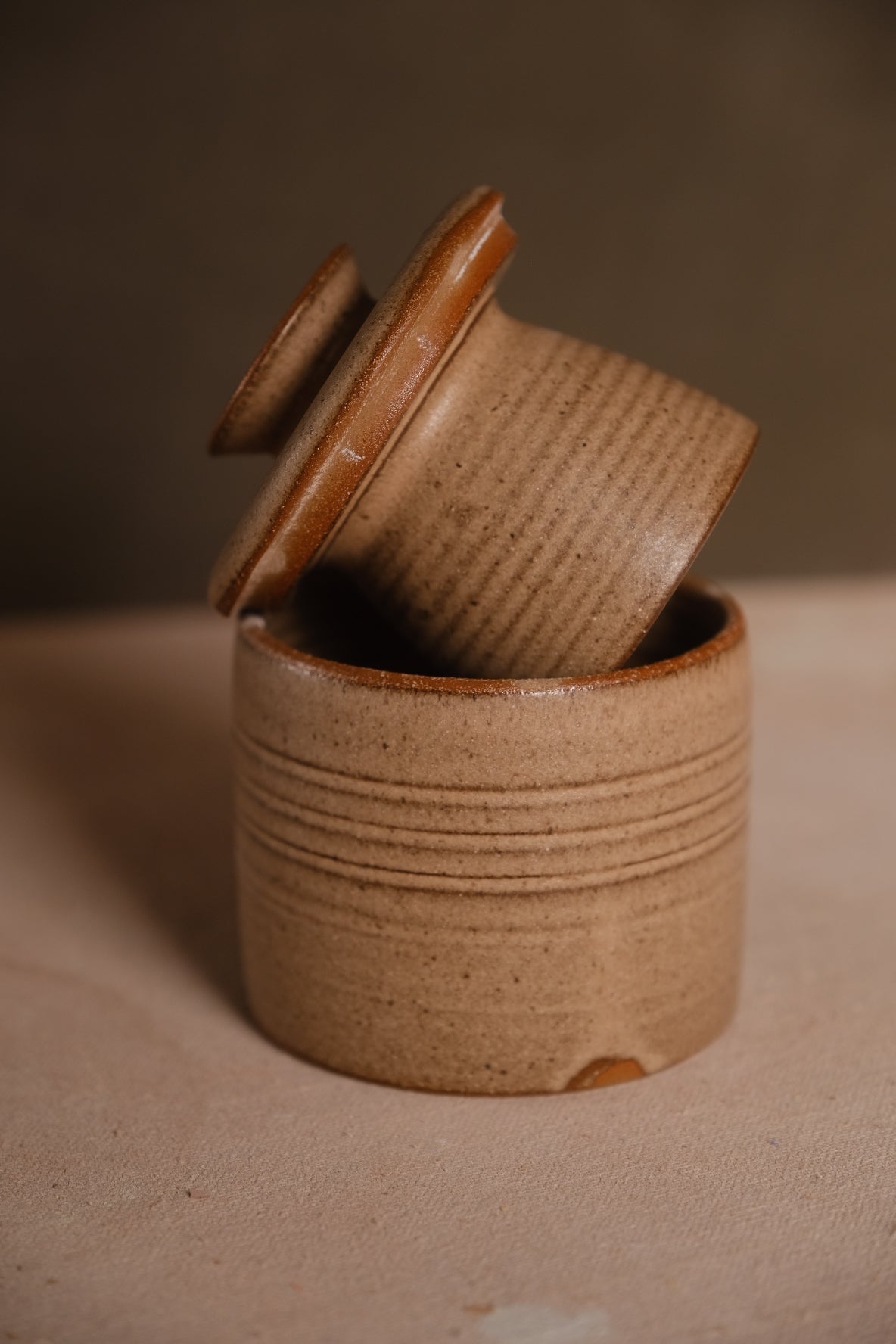 Butterbell keeper featuring ribbed lines across both pieces, lid tilted on base, in speckled tan stoneware with exposed clay.