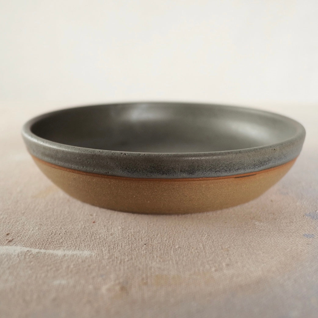 Country Rim Dinner Bowl with dark grey interior glaze and raw stoneware base, side view on canvas.