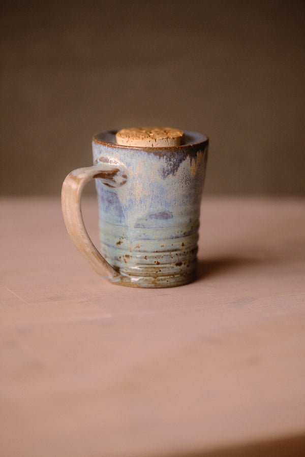 Secret Cenote Traveller Mug standing on a wooden surface, showing its left-facing looped handle, ribbed base, and flush cork.