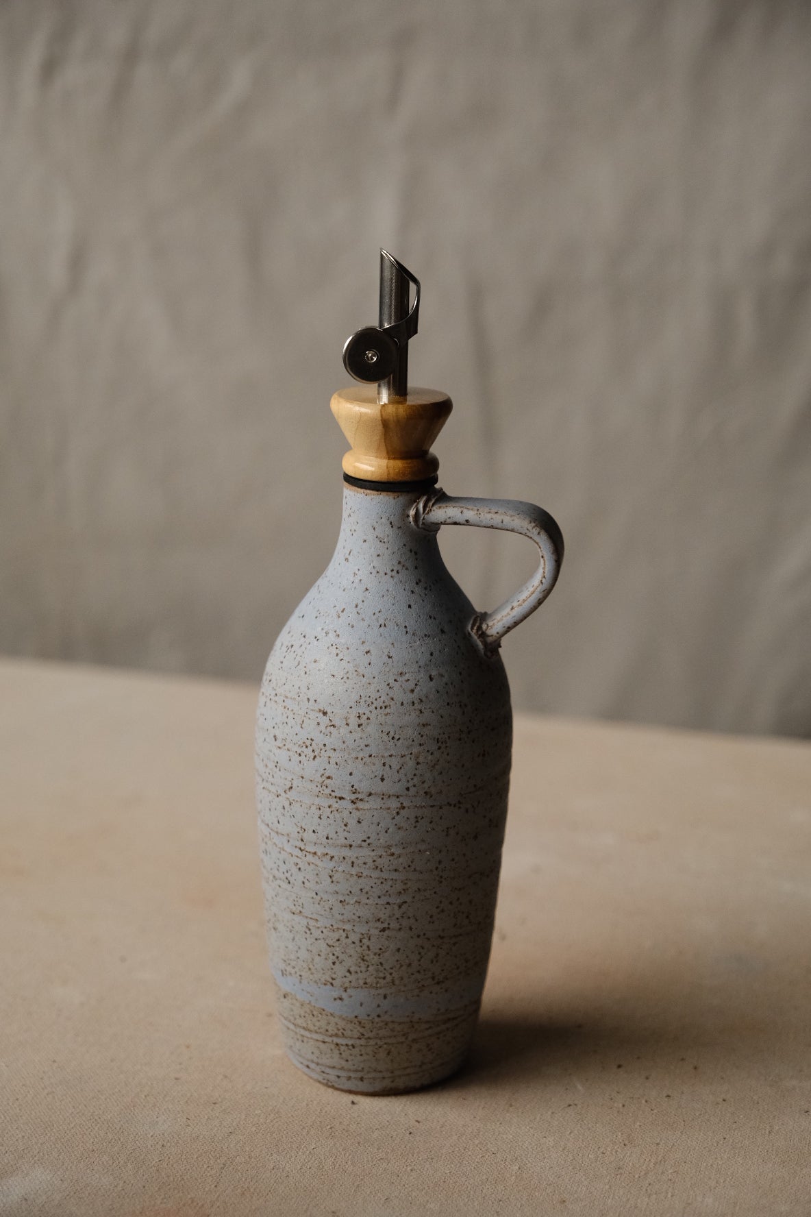 Tejas Oil Bottle in pale blue-grey speckled glaze with fine horizontal rings, side handle, metal spout on a flat surface.