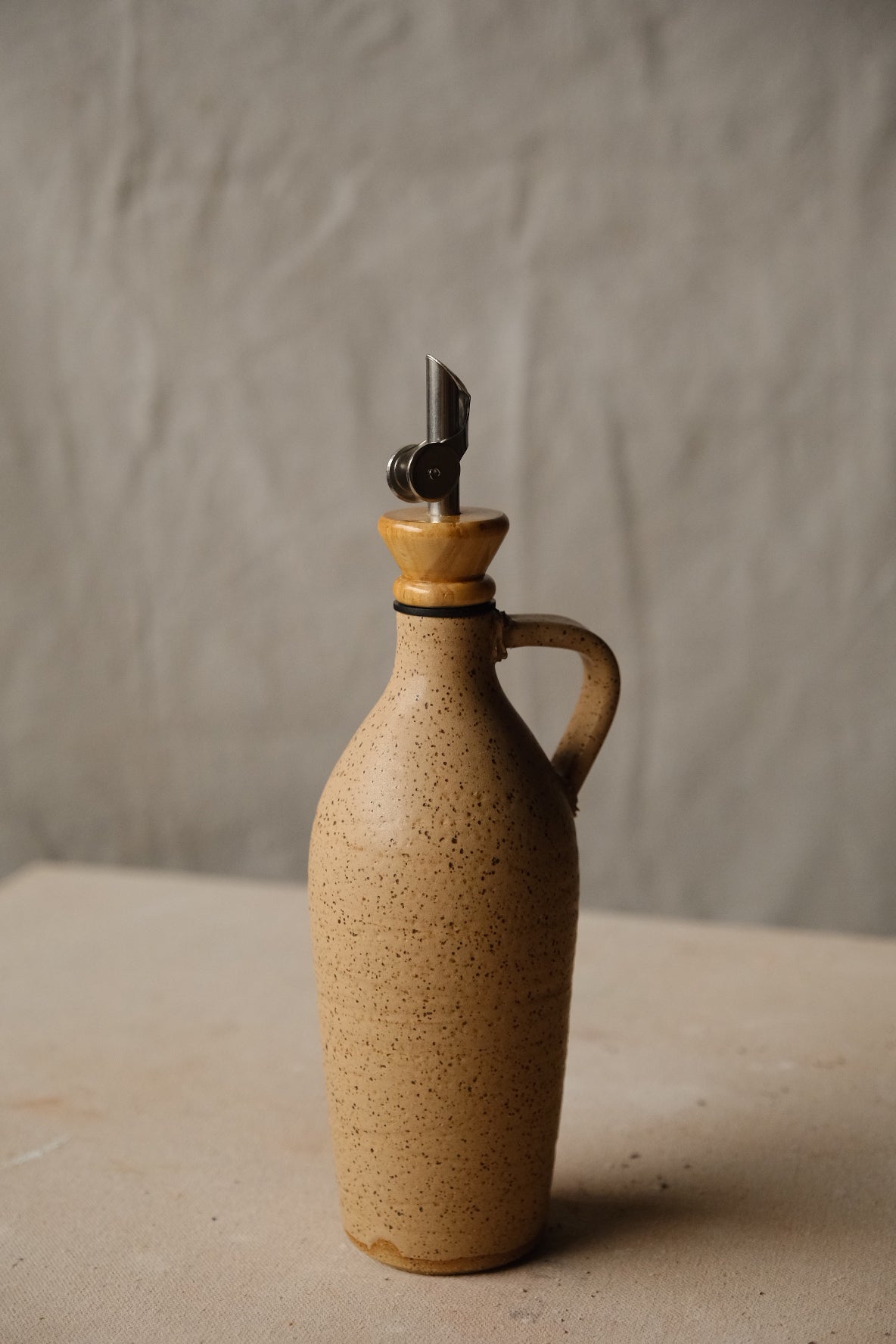 Tejas Oil Bottle in warm tan speckled stoneware, tapered body, loop handle, metal spout with wood collar.