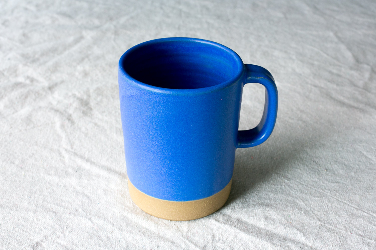 Wander Cappuccino Mug finished in vibrant cobalt blue glaze with side handle and unglazed raw clay foot on linen.