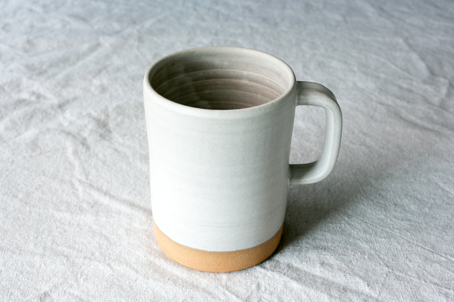 Wander Cappuccino Mug in matte satin white glaze with squared handle and raw clay base resting on linen fabric.
