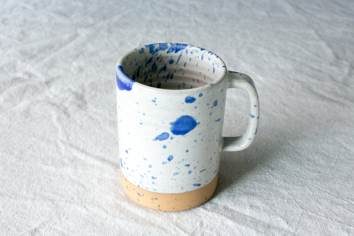 Wander Cappuccino Mug in white with cobalt blue speckles, strap handle, and exposed raw clay base on textured linen.