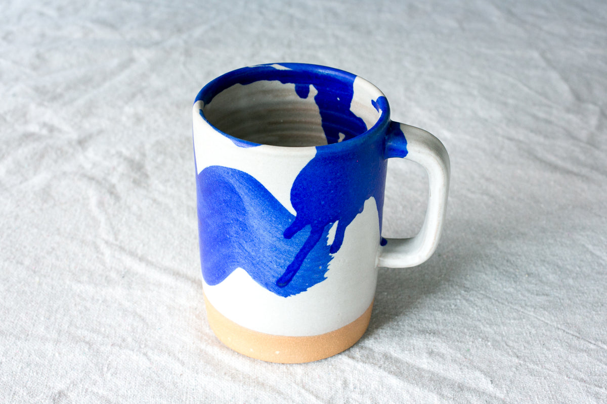 Wander Cappuccino Mug in white glaze with bold blue brushstrokes and splashes, standing on a linen tablecloth.