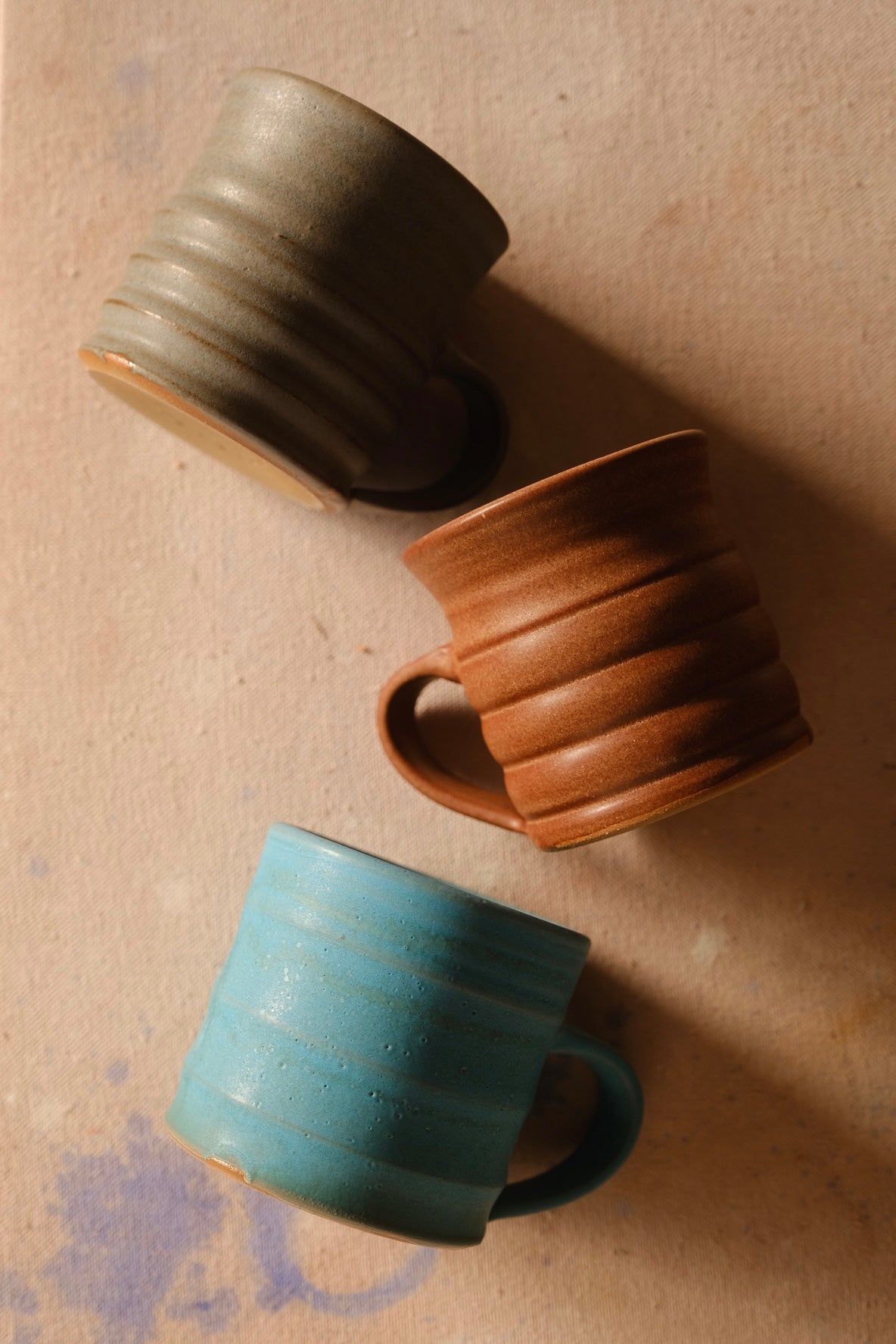 The Big Mug: three ribbed stoneware mugs in different glazes lying on a canvas cloth, seen from above. Hand thrown by Settle Ceramics in Austin, Texas.