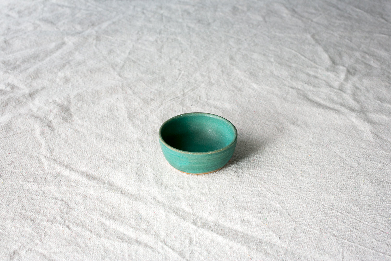 Wander Tiny Bowl in matte teal glaze with an exposed raw clay foot, viewed from above on textured cloth.