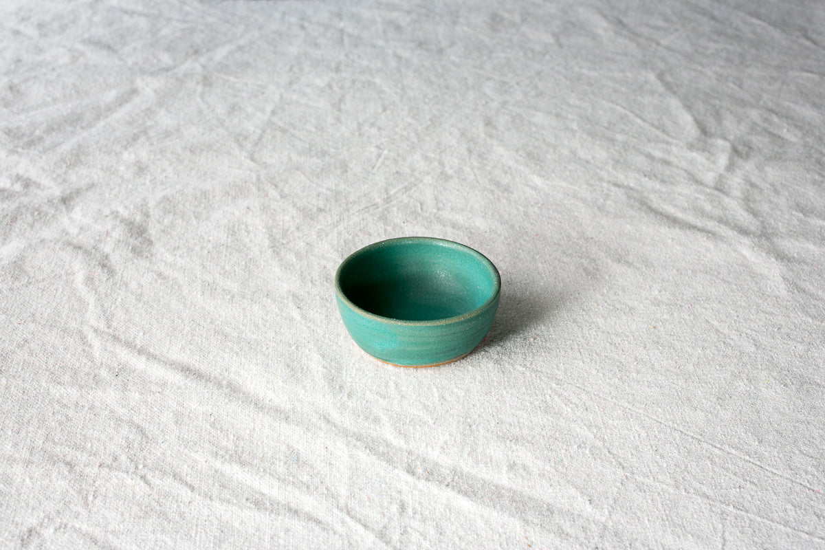Wander Tiny Bowl in matte teal glaze with an exposed raw clay foot, viewed from above on textured cloth.