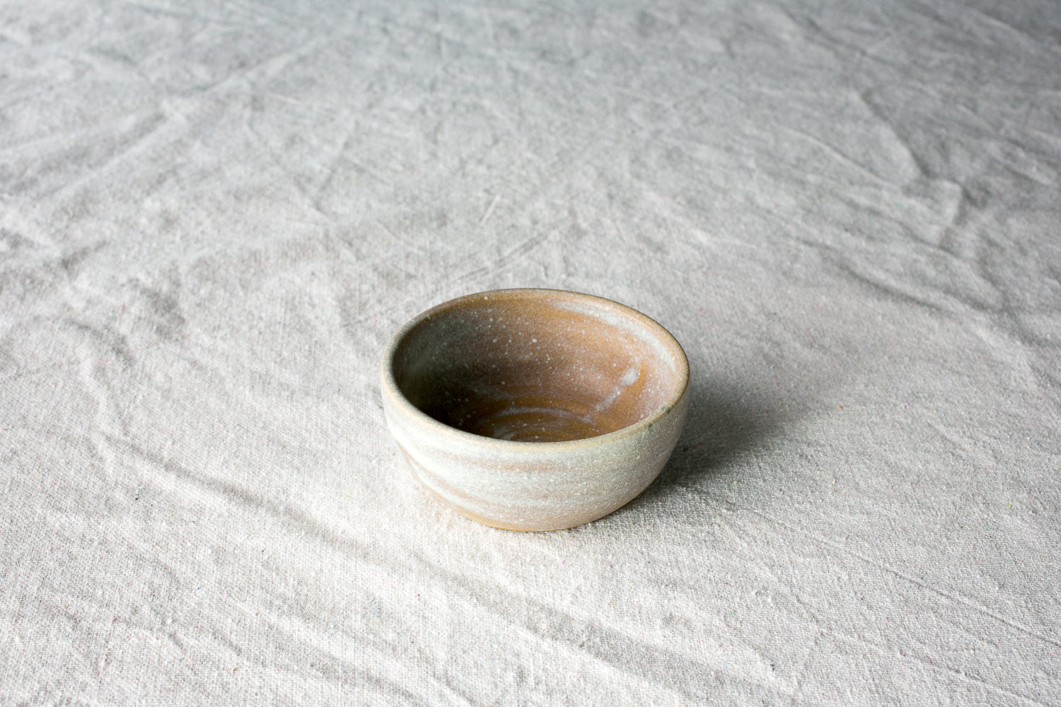 Wander Pinch Bowl, exterior in pale matte glaze with a mottled tan-amber pooled interior, elevated angle on linen.