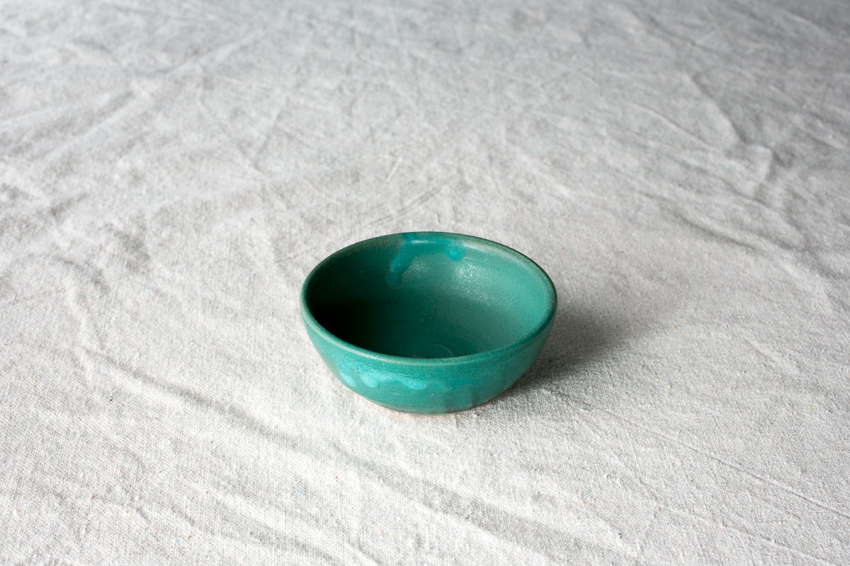 Wander Pinch Bowl in vibrant teal green glaze sitting angled on light woven linen cloth.