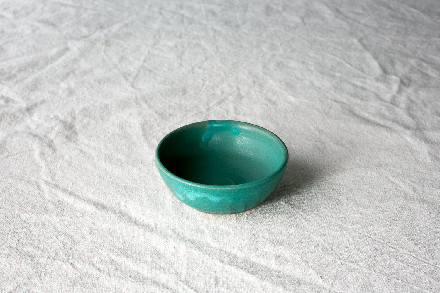 Wander Pinch Bowl in vibrant teal green glaze sitting angled on light woven linen cloth.