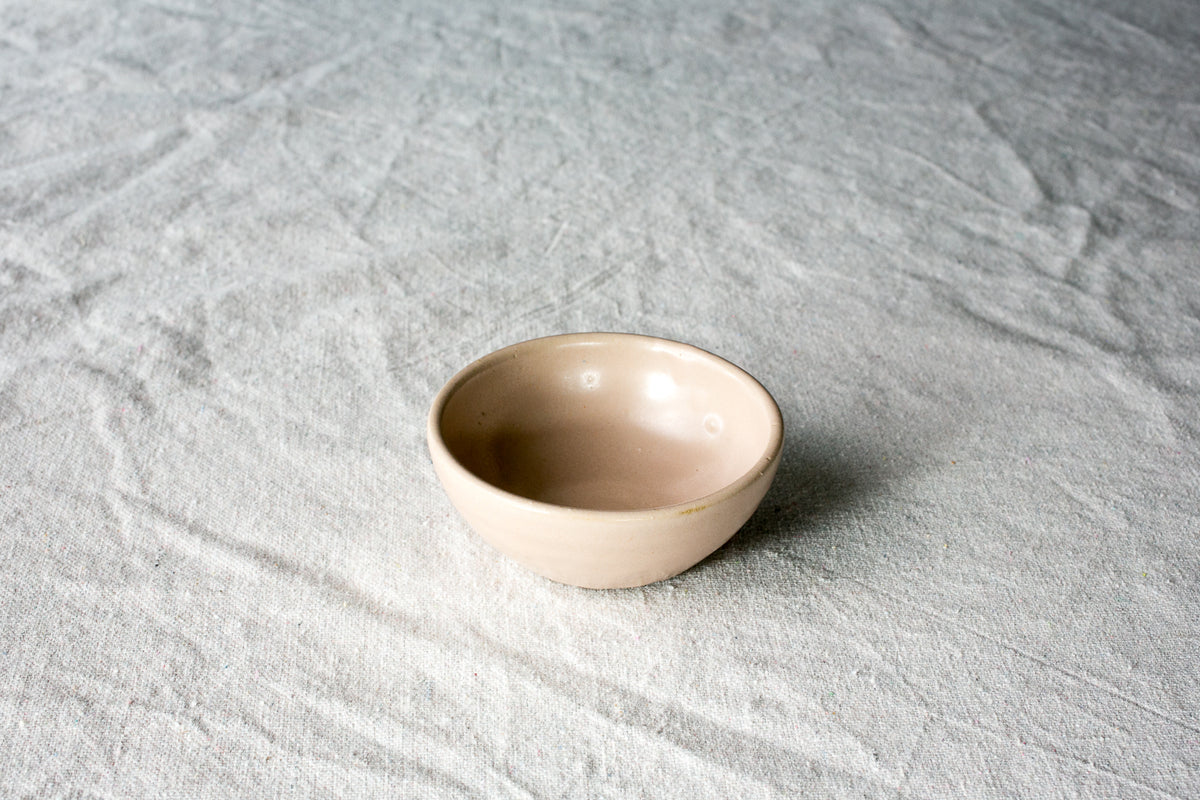 Wander Pinch Bowl in a smooth pale sandy-buff glaze, resting at an angle on neutral linen.