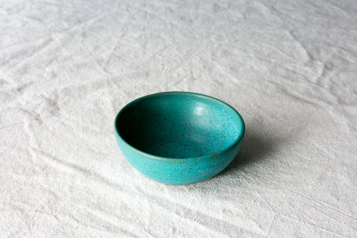 Wander Side Bowl single round dish in teal glaze with subtle speckles resting on textured linen fabric.