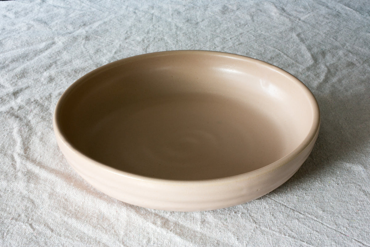 Wander Dinner Bowl in smooth peach-beige glaze, 3/4 high-angle view on textured linen fabric