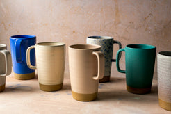 Wander Latte Mug collection, seven mugs in varied glazes lined up on canvas, showing studio stamps.