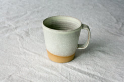 Wander Americano Mug in speckled off-white glaze with strap handle and exposed clay foot on woven fabric.