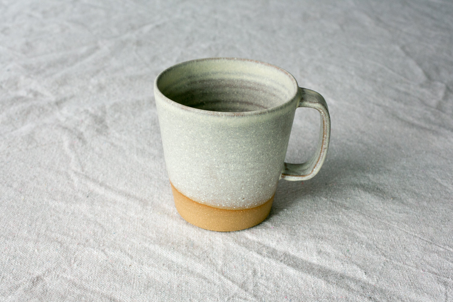Wander Americano Mug in speckled off-white glaze with strap handle and exposed clay foot on woven fabric.