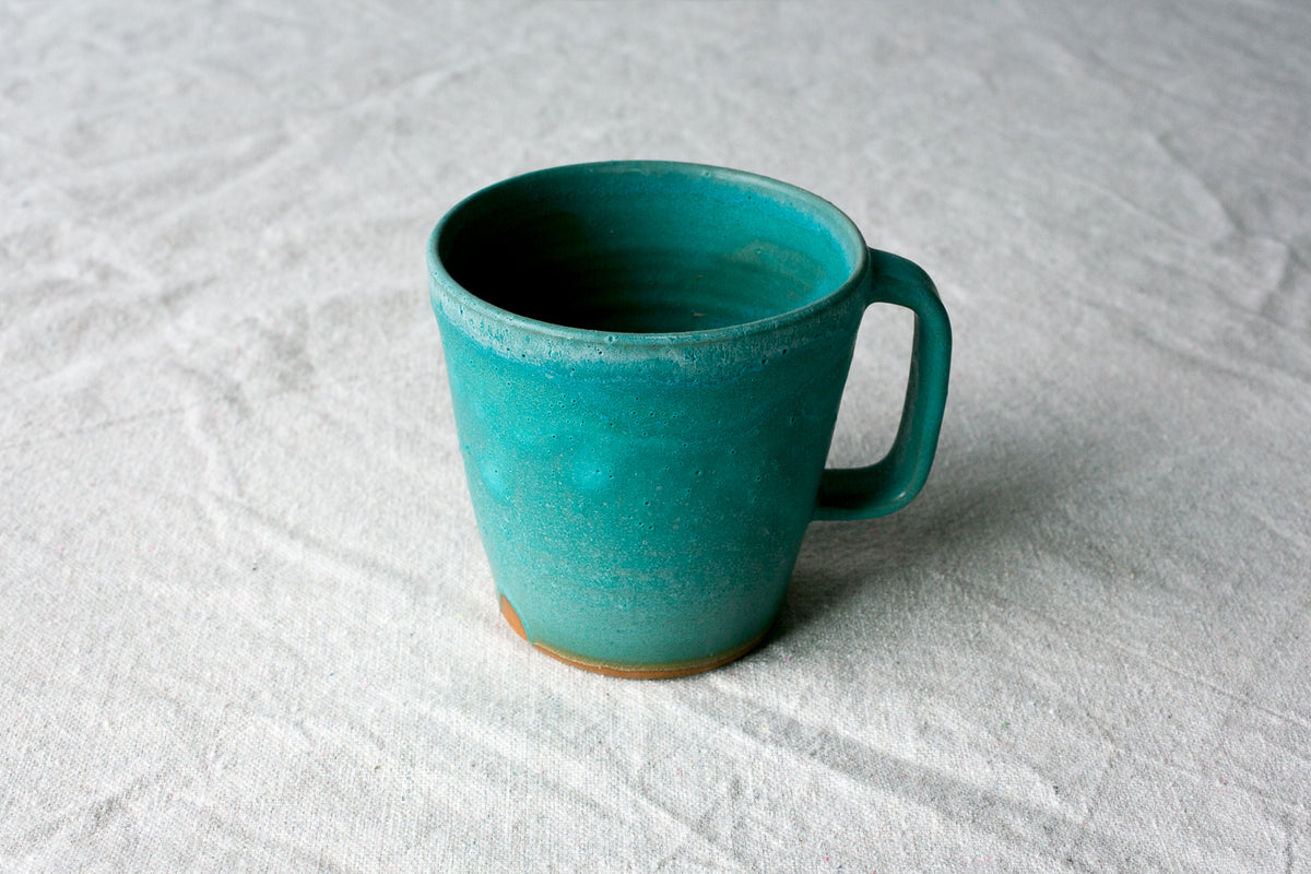 Wander Americano Mug in vibrant turquoise glaze with side handle and unglazed base, resting on light linen.