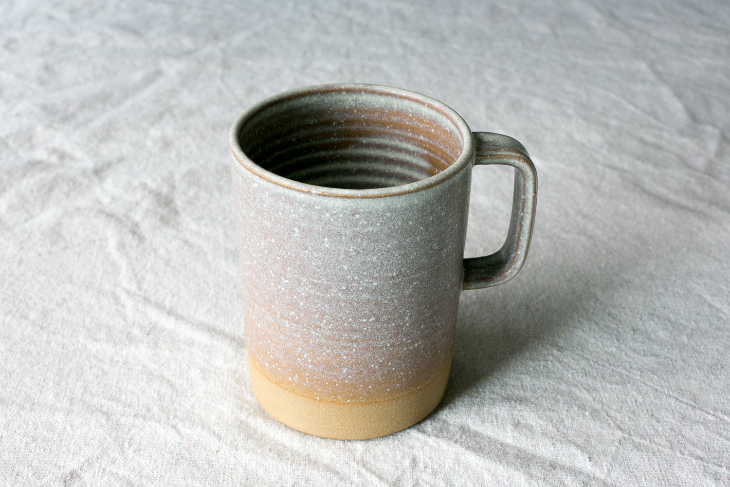 Wander Cappuccino Mug in speckled beige glaze with a square handle and raw exposed clay foot on linen fabric.
