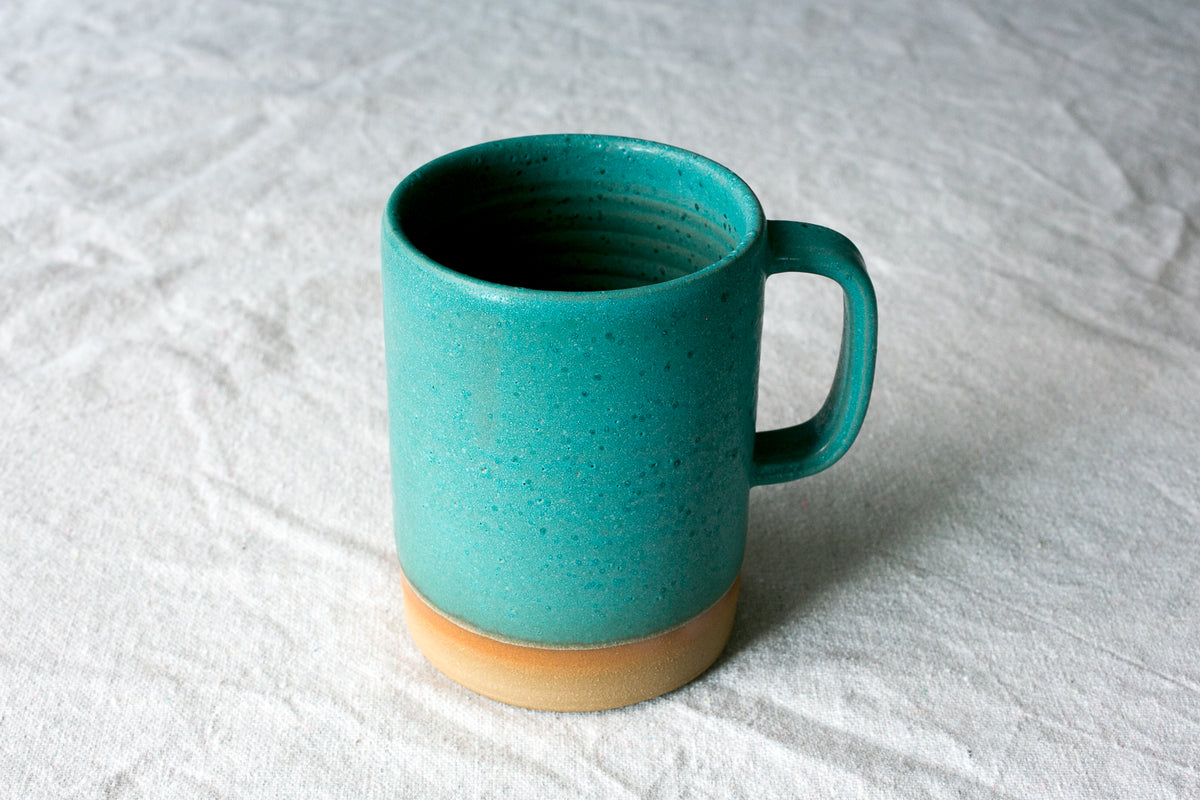 Wander Cappuccino Mug in bright turquoise glaze showing throwing ridges, side handle, and exposed raw clay foot.