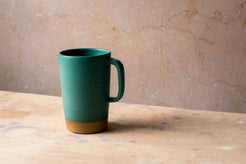 Wander Latte Mug in matte green glaze, side handle view on canvas against mottled wall.