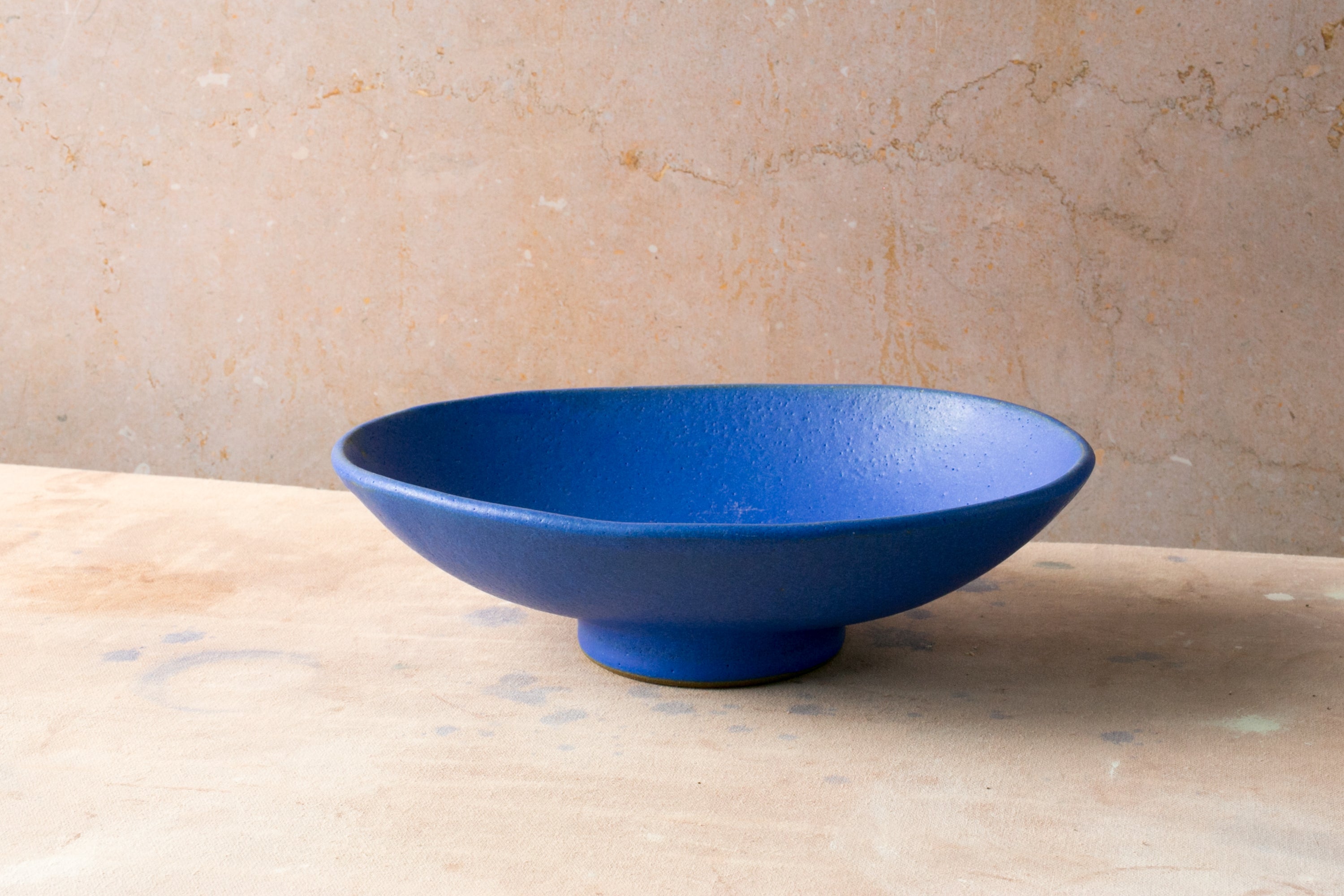 Wander Footed Oval Server in matte cobalt blue glaze on a raised foot, viewed from the front against a warm wall.