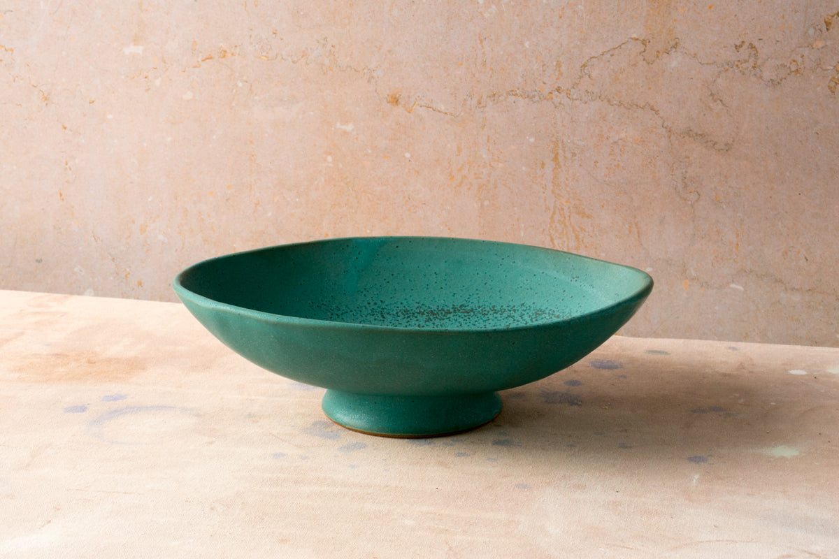 Wander Footed Oval Server in matte teal glaze on an unglazed foot, seen at eye level on a light surface.