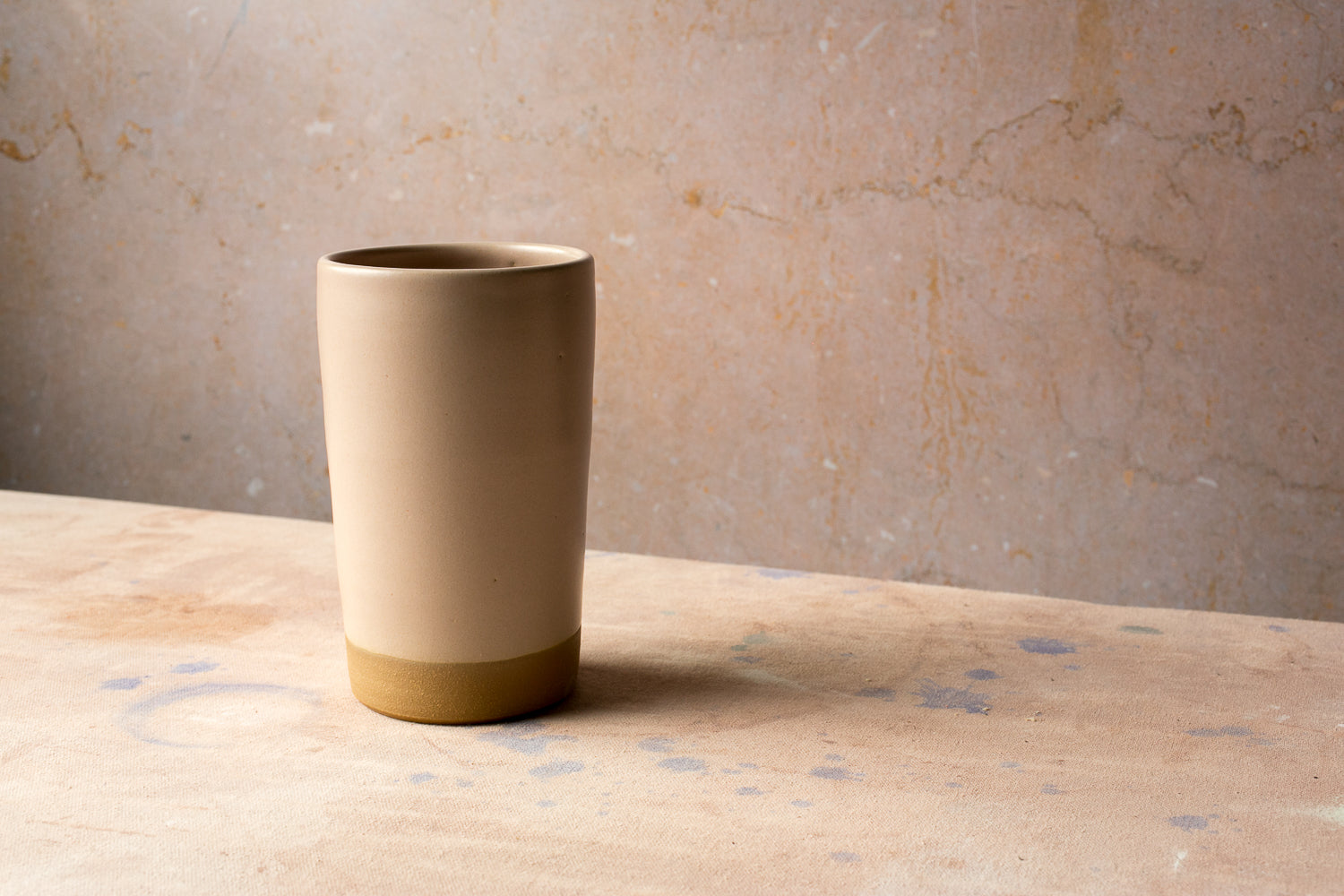 Wander Tall Tumbler in a smooth satin glaze with an unadorned raw clay foot, resting on a tabletop against a wall.