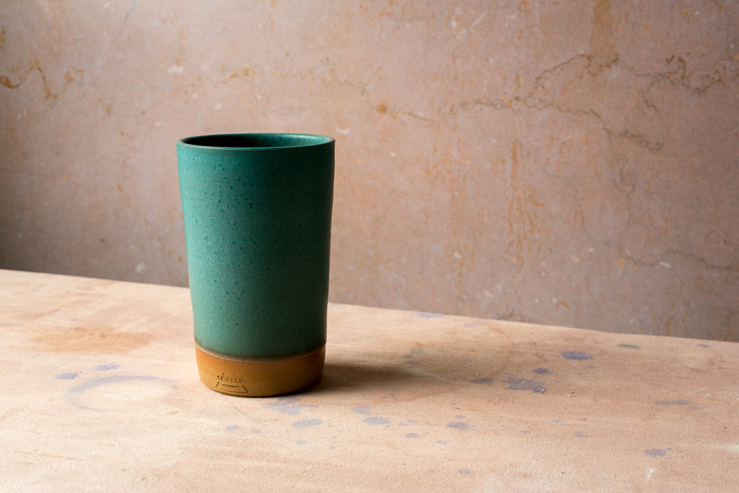 Wander Tall Tumbler in a dark teal glaze, displaying fine speckles and a crisp studio stamp on the raw clay base.