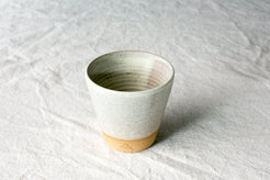 Wander Tumbler with speckled off-white glaze and exposed raw clay foot with maker stamp, sitting on linen fabric.