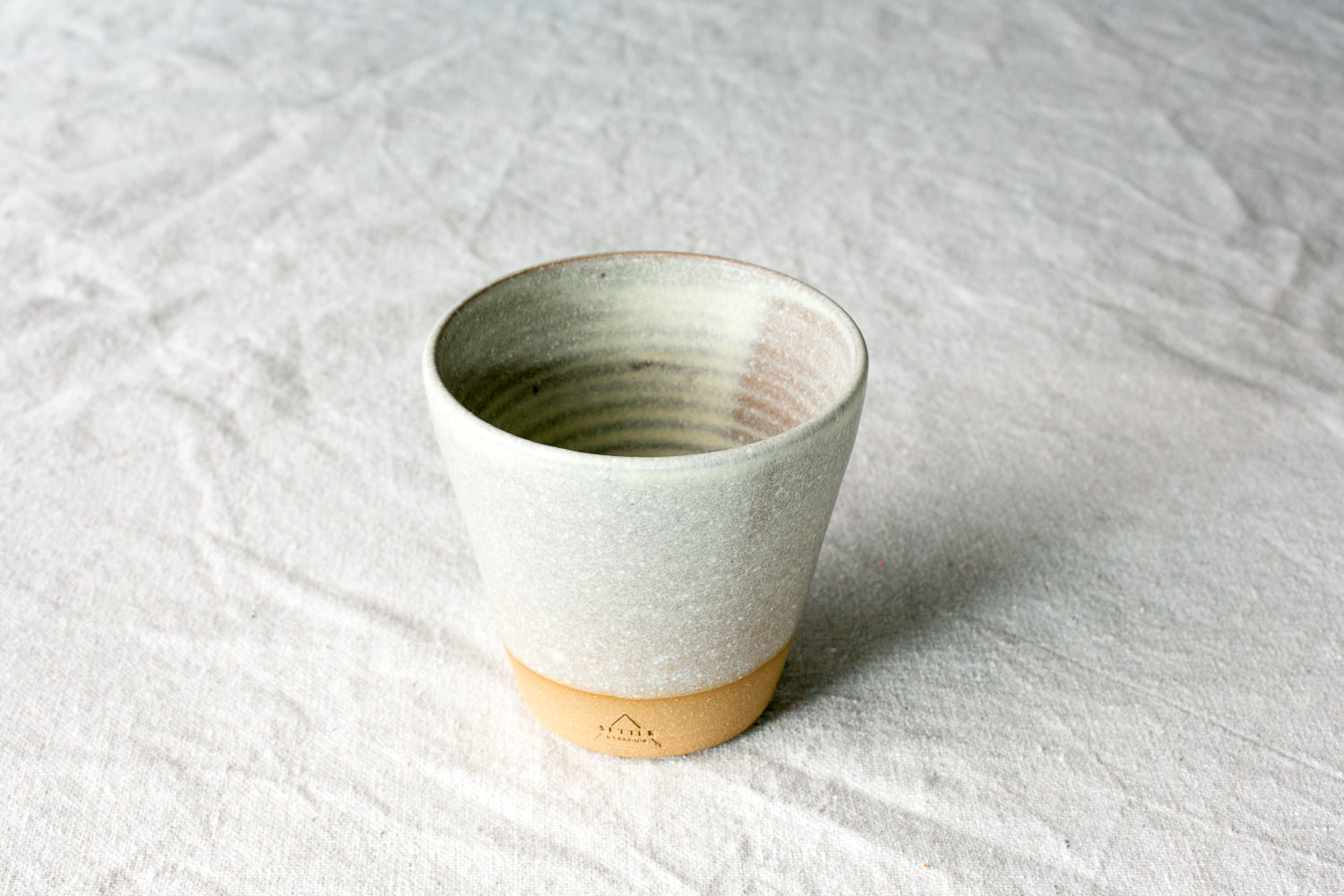 Wander Tumbler with speckled off-white glaze and exposed raw clay foot with maker stamp, sitting on linen fabric.