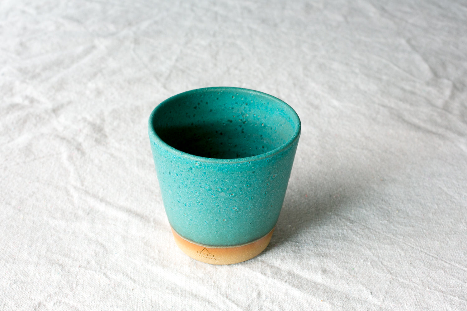 Wander Tumbler with textured turquoise glaze and an exposed raw clay foot with maker stamp, set on linen fabric.