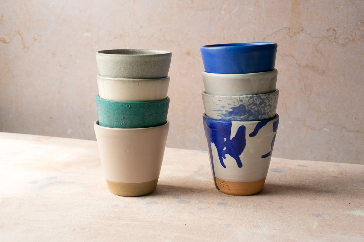 Wander Tumblers stacked in two columns of four, showing varied glazes and exposed clay bases on a warm table.
